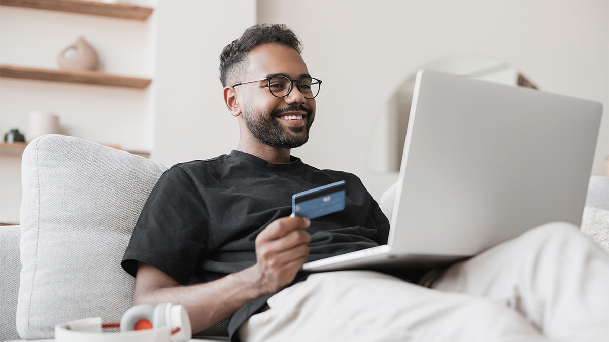 Smiling man on a couch using a laptop and holding a credit card while comparing a credit union credit card to a big-bank option.